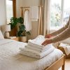 Mujer doblando ropa blanca recién lavada sobre la cama en un dormitorio luminoso, resultado de lavar bien la ropa blanca en la lavadora adecuada, Almacén Electrohogar
